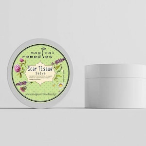 May include: Two white cosmetic jars. One is a round tin with a green label that reads "Magical Remedies Scar Tissue Salve." The label has floral illustrations and the website address. The other jar is a small, cylindrical container.