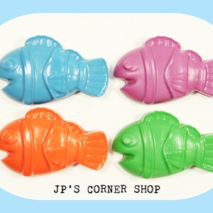 May include: Four colorful fish-shaped crayons in blue, pink, orange, and green.  The crayons are arranged in a square pattern on a white background.  The text "JP'S CORNER SHOP" is printed below the crayons.