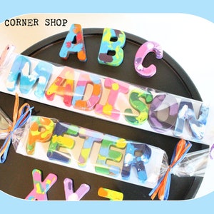 May include: A set of colorful crayon letters spelling out the names "Madison", "Peter", and "ABC" on a black surface. The letters are packaged in clear cellophane bags with colorful ribbons.