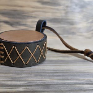 May include: A handcrafted leather drum-shaped item with a brown top and dark green sides. The sides are stitched with a crisscross pattern using tan thread. A leather strap is attached.
