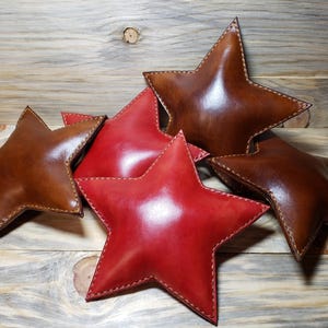 Pode incluir: Cinco almofadas em forma de estrela, duas de couro marrom e três de couro vermelho. As almofadas estão empilhadas em uma superfície de madeira.