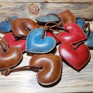Puede incluir: Un grupo de adornos de cuero en forma de corazón en colores marrón, rojo y azul. Los corazones tienen un borde cosido y están encordados.