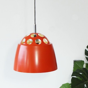 May include: A bright red pendant light with a rounded, bell-like form. The top section has evenly spaced circular cutouts. The light hangs from a black cord and a silver-toned metal rod. The design is a retro style.