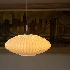 Könnte beinhalten: Eine weiße, gerippte, untertassenförmige Glas-Pendelleuchte, die an einem dünnen Kabel hängt. Die Lampe hat eine glatte, abgerundete Silhouette mit vertikalen Rillen, die von der Mitte ausgehen, und ist mit einer Chromkappe und einem weißen Stab versehen.