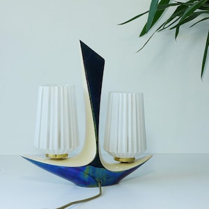 Può includere: Una lampada da tavolo a due luci con una base scultorea e curva caratterizzata da una finitura lucida blu scuro e verde. Due paralumi cilindrici in vetro bianco con coste verticali sono montati su basi circolari color ottone.