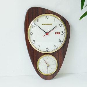 Può includere: Un orologio da parete a forma di goccia con finitura in legno scuro. La sezione superiore presenta un quadrante bianco con numeri neri, bordo dorato e la scritta Morez-Besancon. Un barometro più piccolo con le scritte Variable, Pluie e Beau si trova sotto.
