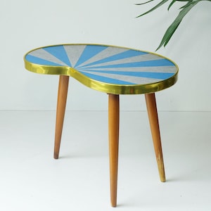 May include: A mid-century modern side table with a kidney-shaped top. The tabletop features a blue and silver sunburst design, framed by a brass edge. It stands on three tapered wooden legs, creating a stylish accent piece.