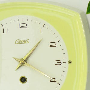 May include: Close-up of a vintage Comet clock with a pale yellow square frame. The clock face is white with black numerals and gold hands. The word "Comet" is printed in cursive. Green leaves are visible in the background.