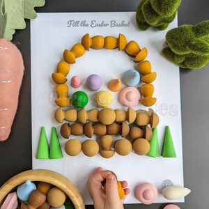Może przedstawiać: Biała kartka z nadrukowanym tekstem "Fill the Easter Basket". Kartka jest otoczona różnymi kolorowymi drewnianymi kształtami, w tym stożkami, kulami i walcami. Kształty są ułożone w okrąg wokół tekstu.