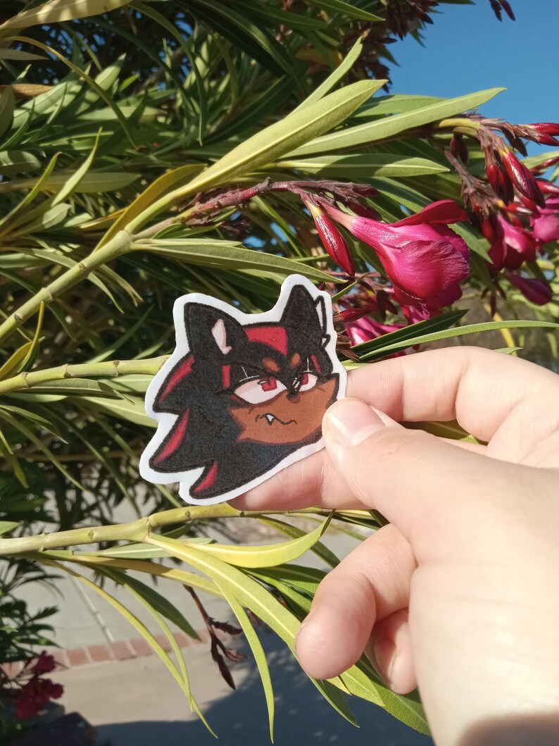 K&ouml;nnte beinhalten: Ein schwarzer und roter Comic-Charakter mit einem rot-schwarz gestreiften Shirt und wei&szlig;em Hintergrund. Der Charakter hat einen schelmischen Ausdruck und h&auml;lt eine rote Blume.