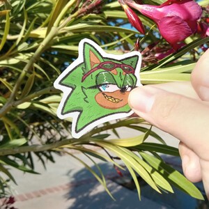 K&ouml;nnte beinhalten: Ein gr&uuml;ner Comic-Charakter mit einem roten Bandana, Sonnenbrille und einem goldenen Zahn. Der Charakter h&auml;lt einen rot-wei&szlig; gestreiften Lollipop.
