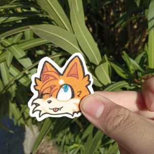 K&ouml;nnte beinhalten: Ein Aufkleber mit einem Comic-Fuchs mit orangefarbenem Fell, blauen Augen und einer herausgestreckten rosa Zunge. Der Fuchs schaut nach rechts und hat einen schelmischen Ausdruck.