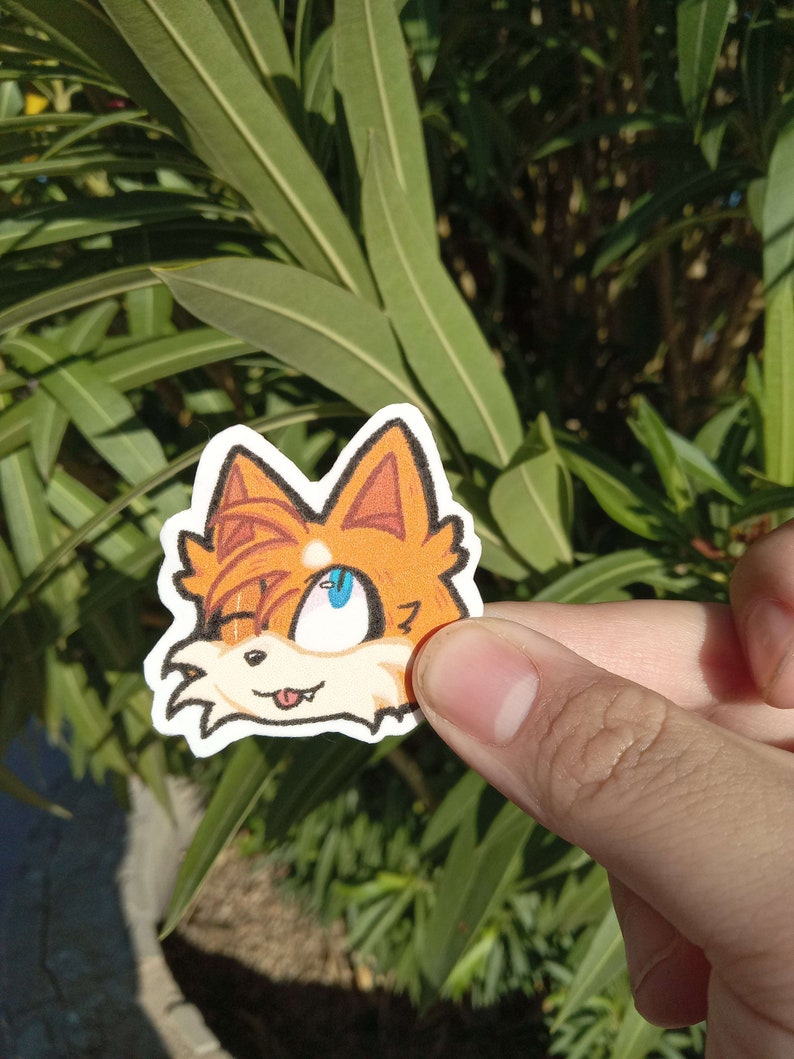 K&ouml;nnte beinhalten: Ein Aufkleber mit einem Comic-Fuchs mit orangefarbenem Fell, blauen Augen und einer herausgestreckten rosa Zunge. Der Fuchs schaut nach rechts und hat einen schelmischen Ausdruck.