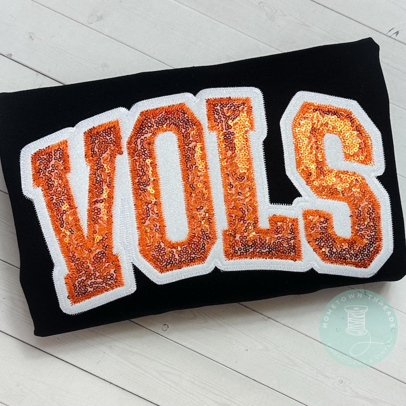 May include: Black fabric with the word "VOLS" in orange sequined letters outlined in white. The letters are in a bold, collegiate-style font. A small logo with a spool of thread is in the bottom right corner.
