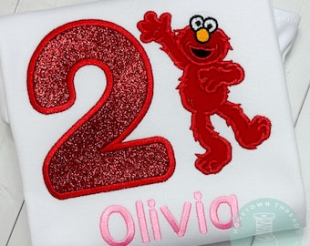 May include: White t-shirt with a large red glittery number 2 and a red cartoon character. The name "Olivia" is embroidered in pink script below the number. The character is waving.