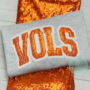 May include: A gray sweatshirt with the word "VOLS" in orange sequined letters. The sweatshirt is displayed on a background of orange sequined fabric. The design is a collegiate or sports-themed item.