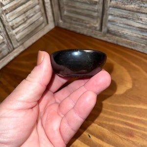 May include: A small, black, polished bowl with a rounded shape. The bowl is held in a hand, showcasing its smooth, reflective surface. The bowl's dark color contrasts with the wooden background.