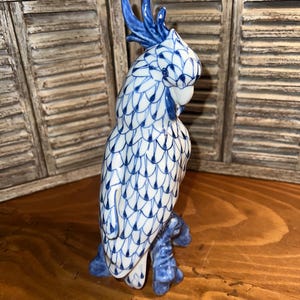 May include: A ceramic figurine of a cockatoo, predominantly white with intricate blue patterns. The bird is perched on a blue branch, with a detailed crest and beak. The figurine is a decorative item.