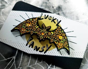 Può includere: Uno spillo in smalto nero con un design floreale giallo e arancione a forma di pipistrello. Lo spillo dice "Lucky Pansy" sul retro.