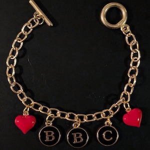 May include: Gold-tone chain bracelet with a toggle clasp. The bracelet features three black circular charms with the letters "B", "B", and "C", and two red heart charms. The bracelet is displayed on a black surface.