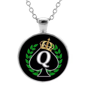 May include: A silver necklace with a round pendant featuring a black spade with a white queen letter and a gold crown on top. The spade is surrounded by green laurel leaves on a black background.