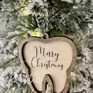 May include: A wooden tooth-shaped Christmas ornament with the text "Merry Christmas" engraved on it. The ornament is hanging on a snowy Christmas tree.