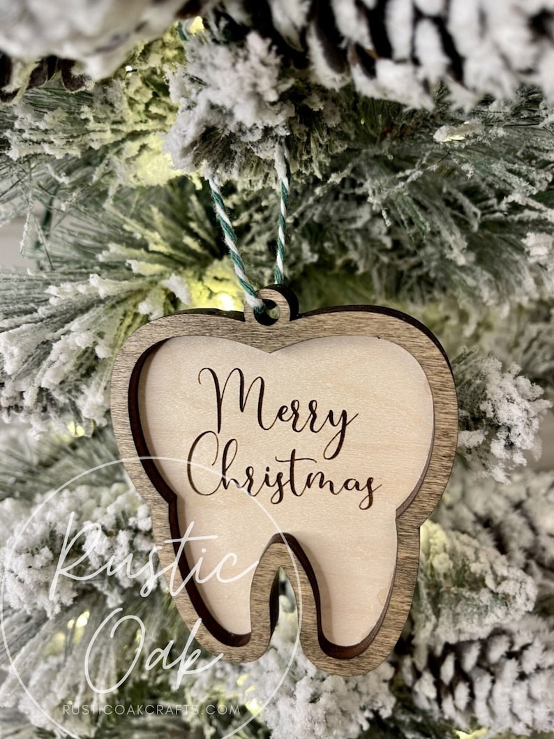 May include: A wooden tooth-shaped Christmas ornament with the text "Merry Christmas" engraved on it. The ornament is hanging on a snowy Christmas tree.