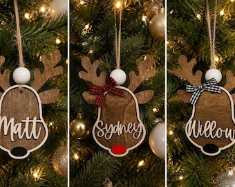 May include: Three wooden reindeer ornaments featuring antlers, a white bead, and a jute string. Each ornament displays a name in white script: Matt, Sydney, or Willow. The reindeer have small bows and colored noses in black or red.