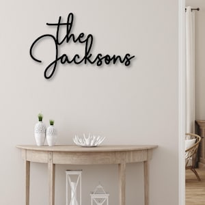 May include: Black cursive metal sign that reads "the Jacksons" mounted on a light-colored wall. The sign is above a wooden console table with decorative items, including vases and lanterns. The room has a neutral color palette.