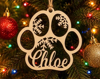 May include: A light-colored wooden ornament shaped like a paw print, featuring cut-out snowflake designs and the name Chloe in cursive script. The ornament is suspended by a thin, natural-colored twine loop for hanging on a tree branch.