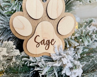 May include: A wooden paw print ornament featuring a dark brown base layer and lighter wood-toned accents for the pads. The central pad is engraved with the name Sage in a cursive script. A thin twine loop is attached at the top for hanging.