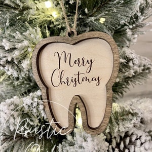 May include: A wooden tooth-shaped Christmas ornament with the text "Merry Christmas" engraved on it. The ornament is hanging on a snowy Christmas tree.