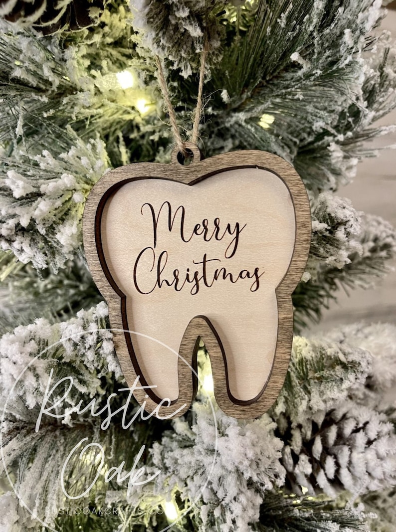 May include: A wooden tooth-shaped Christmas ornament with the text "Merry Christmas" engraved on it. The ornament is hanging on a snowy Christmas tree.