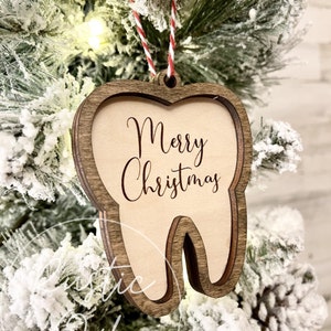 May include: A wooden tooth-shaped Christmas ornament with the words "Merry Christmas" inscribed on it. The ornament is hanging on a Christmas tree.