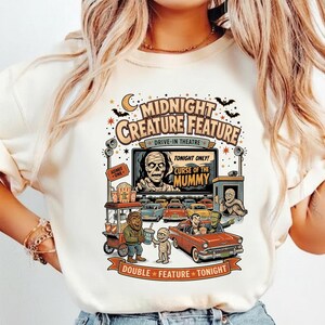 Puede incluir: Una camiseta de color crema con un gráfico de un autocine con monstruos. El texto dice Midnight Creature Feature, Drive-In Theatre, Tonight Only, Curse of the Mummy, Admit One y Double Feature Tonight.