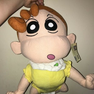 May include: A large plush toy of a baby with brown hair, wearing a yellow onesie with a white collar and a green leaf design. The toy has large black eyes and a surprised expression.