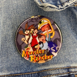 May include: A round button featuring characters from the animated series "Monster Rancher" is pinned to a denim jacket. The button has a dark background with cartoon characters in various colors, and the show's title is in orange and yellow.