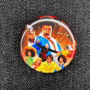 May include: A round button featuring a colorful image of a man with a mustache holding a gun, surrounded by other people. The button has a black background and a red border.