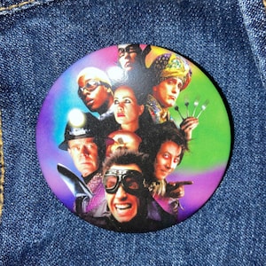 May include: A colorful button featuring a group of people with various expressions. The button has a rainbow background with a green and purple gradient. The people are wearing different outfits, including a hat with a feather, a helmet, and goggles.