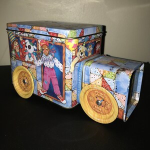 May include: A blue and white tin toy truck with a patchwork design. The truck has a clown and a dog on the side. The truck has two large yellow wheels with a button design.