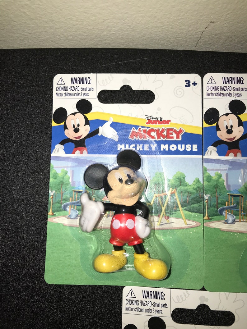 Peut inclure: Une figurine en plastique de Mickey Mouse dans un emballage en plastique transparent. La figurine porte un short rouge avec deux cercles blancs, des chaussures noires et des gants blancs. L'arri&egrave;re-plan est un parc vert avec un toboggan et une balan&ccedil;oire. L'emballage porte une &eacute;tiquette d'avertissement indiquant "RISQUE D'&Eacute;TOUFFEMENT - Petites pi&egrave;ces. Ne convient pas aux enfants de moins de 3 ans."