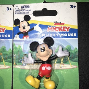 Peut inclure: Une figurine en plastique de Mickey Mouse dans un emballage en plastique transparent avec un fond vert, bleu et jaune. L'emballage porte les mots "Disney Junior Mickey Mickey Mouse". L'emballage comporte &eacute;galement une &eacute;tiquette d'avertissement indiquant "Risque d'&eacute;touffement - Petites pi&egrave;ces. Ne convient pas aux enfants de moins de 3 ans."