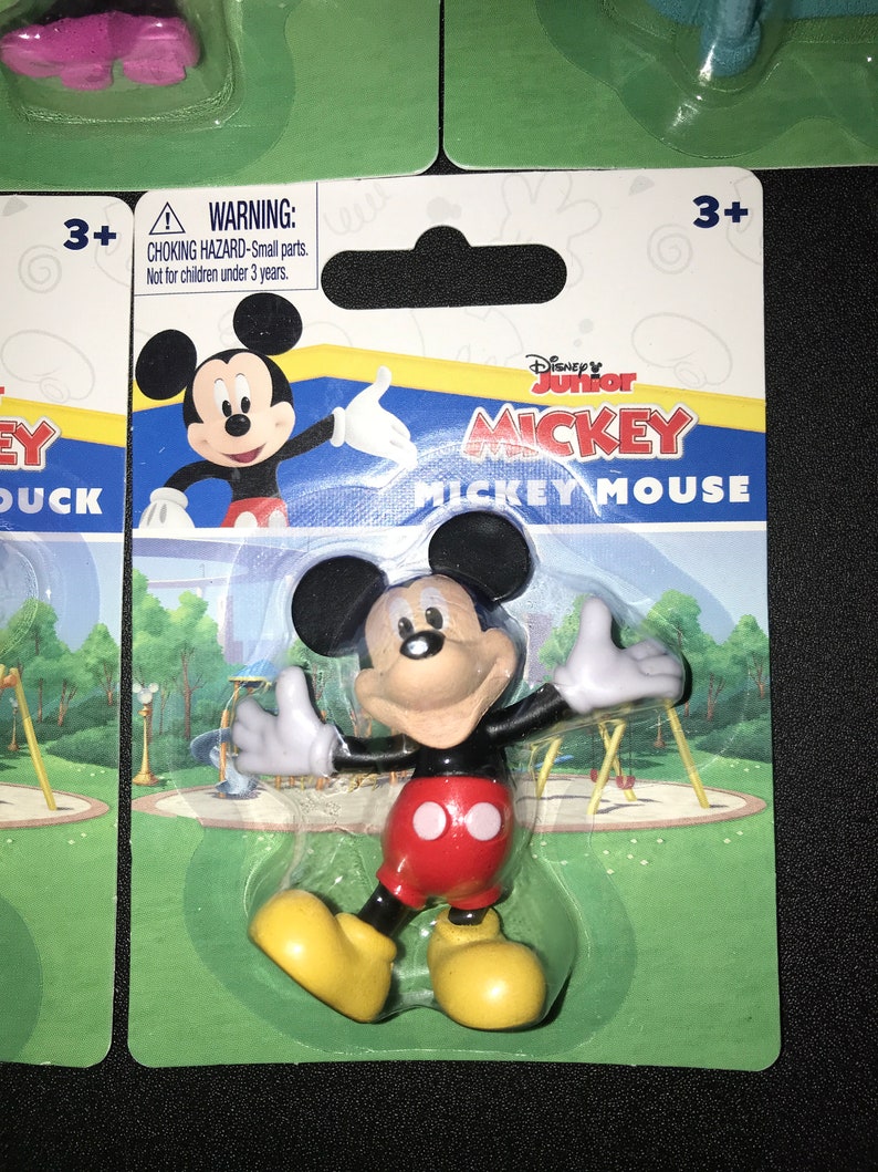 Peut inclure: Une figurine en plastique de Mickey Mouse dans un emballage en plastique transparent avec un fond vert, bleu et jaune. L'emballage porte les mots "Disney Junior Mickey Mickey Mouse". L'emballage comporte &eacute;galement une &eacute;tiquette d'avertissement indiquant "Risque d'&eacute;touffement - Petites pi&egrave;ces. Ne convient pas aux enfants de moins de 3 ans."