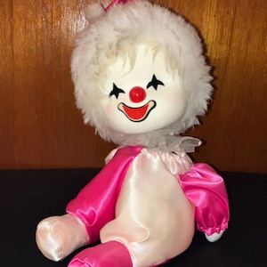 May include: A vintage clown doll with a plush white head and a smiling face with black tear-shaped markings. The doll wears a pink and white satin outfit with a ruffled collar. The doll is sitting down.
