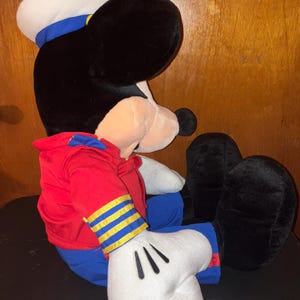 May include: A large plush toy of a cartoon mouse character, dressed in a sailor outfit. The toy has a black head and ears, a white sailor hat with a blue band, a red jacket, blue trousers, and white gloves. The toy is in a seated position.