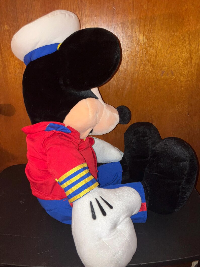 May include: A large plush toy of a cartoon mouse character, dressed in a sailor outfit. The toy has a black head and ears, a white sailor hat with a blue band, a red jacket, blue trousers, and white gloves. The toy is in a seated position.