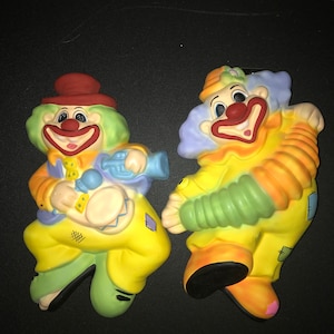 May include: Two ceramic clown figurines, one playing a drum and the other with a green and orange striped sleeve. Both clowns have red noses, yellow and orange clothing, and a whimsical expression.
