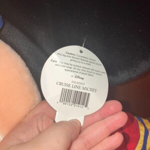 May include: A white tag with the text "CRUISE LINE MICKEY" and a barcode is held in a hand. The tag also includes care instructions and a warning about removing fasteners. The tag is attached to a plush toy.