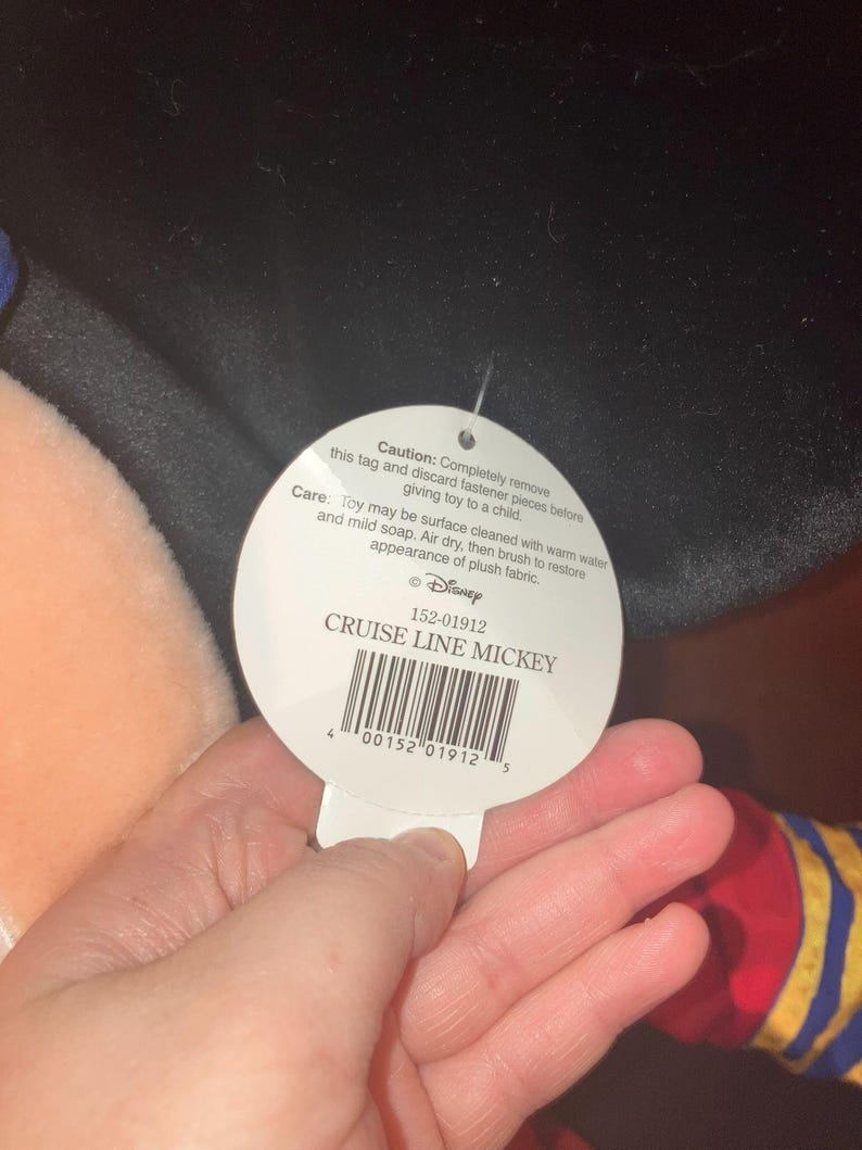 May include: A white tag with the text "CRUISE LINE MICKEY" and a barcode is held in a hand. The tag also includes care instructions and a warning about removing fasteners. The tag is attached to a plush toy.
