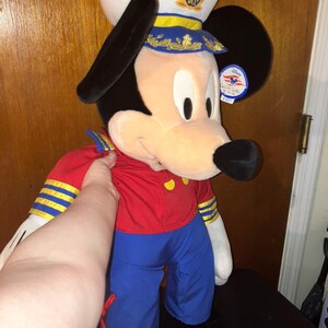 May include: A large plush Mickey Mouse doll wearing a white captain's hat with gold accents and a blue and gold band. Mickey is dressed in a red jacket with gold buttons and blue trousers. A Disney Cruise Line tag is attached.